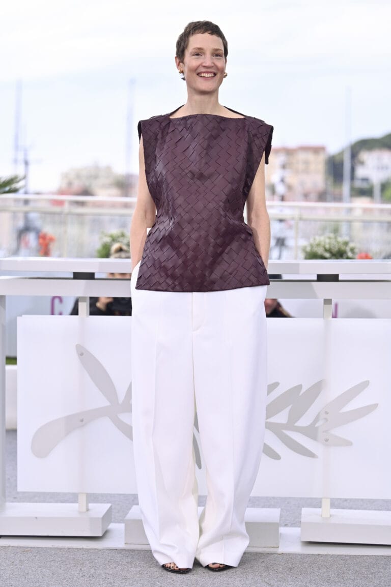 Vicky Krieps: Wears a purple braided-texture blouse and wide-cut white pants, creating…