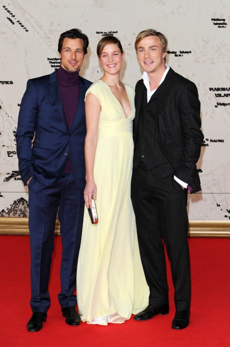 Vicky Krieps: Wears a long light yellow dress with a V-neckline that…