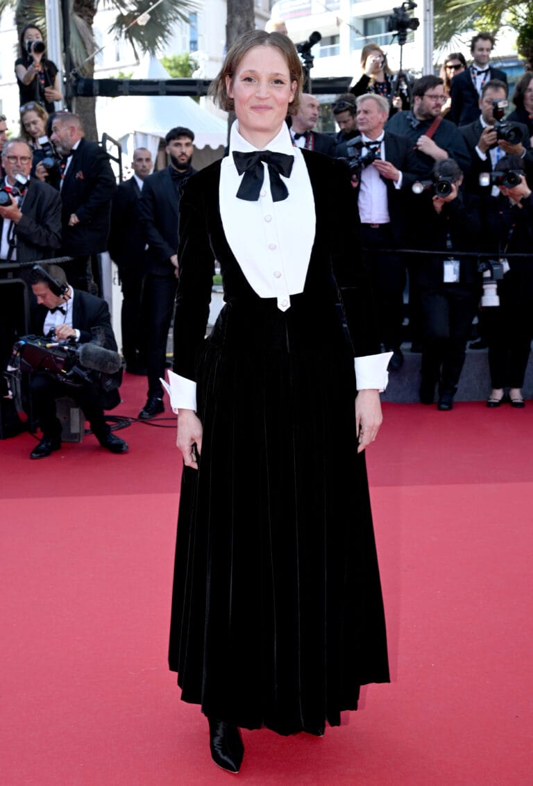 Vicky Krieps: Wears an elegant black velvet dress with a distinctive white collar and a…