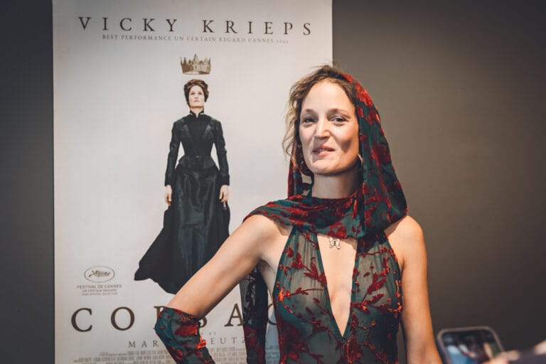 Vicky Krieps: Wears a sheer dress with red floral details that fits…