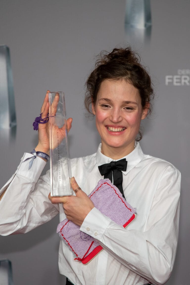 Vicky Krieps: Holds an award in one hand and a cloth bag in the other,…
