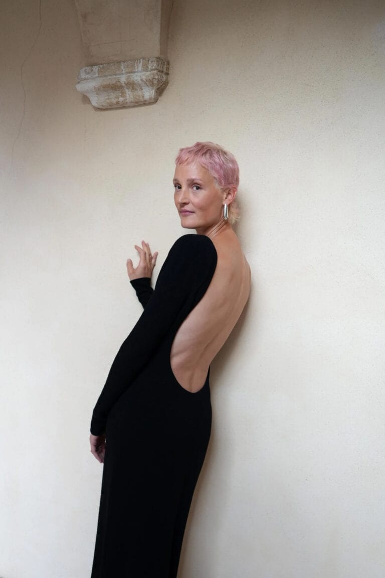 Vicky Krieps: Wears a long black dress with a deep open back, which…