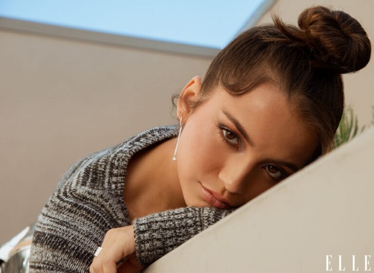 Isabela Merced: Wears a soft-textured gray sweater, resting on a slanted surface with a…