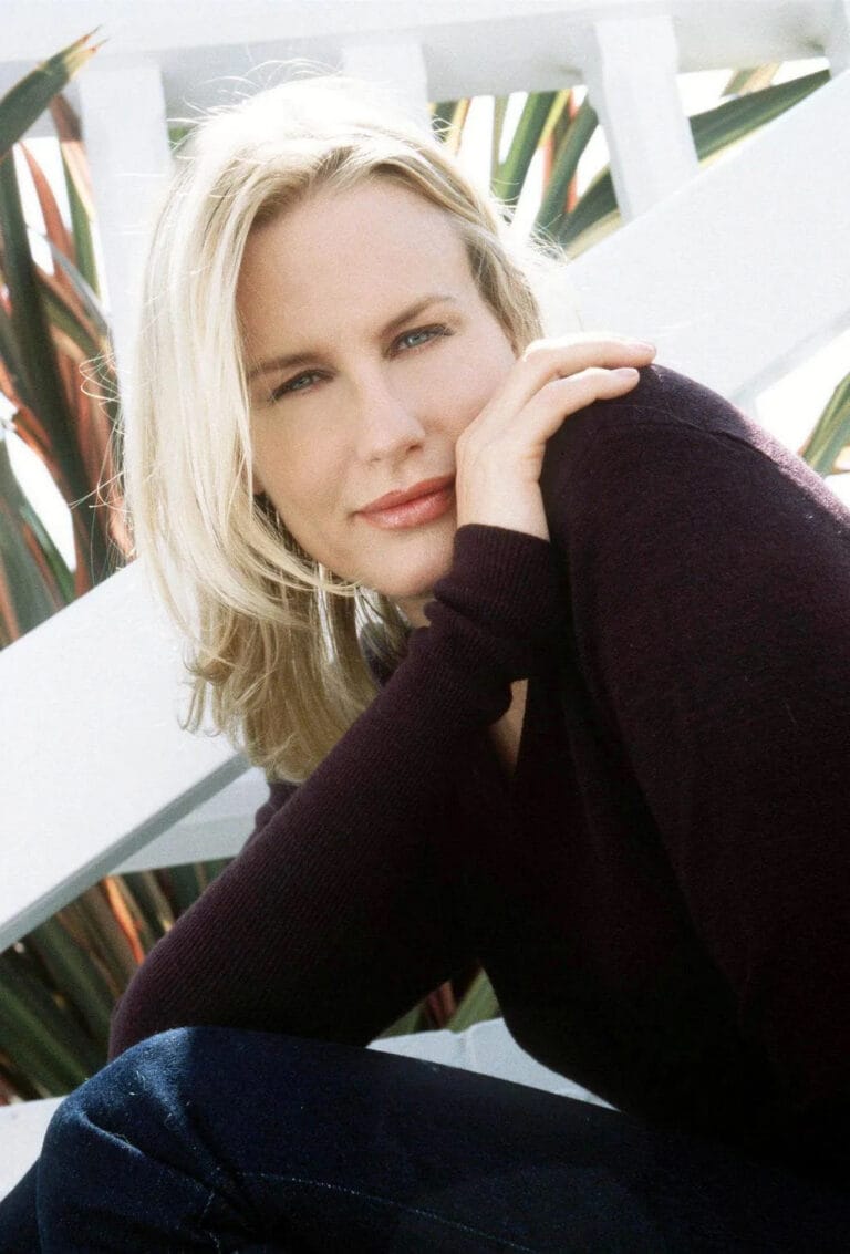 Daryl Hannah: Wears a dark long-sleeve sweater, leaning with one arm on a surface…