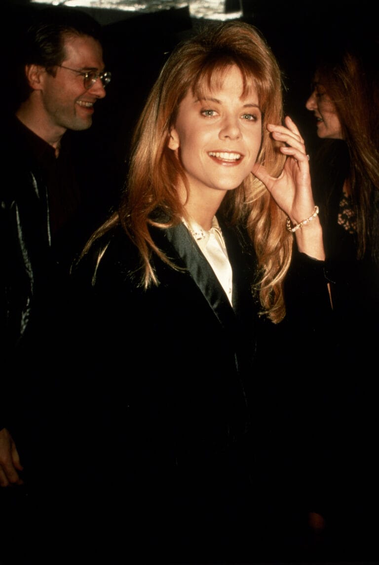 Meg Ryan: Wears an elegant black coat over a white satin top, smiling while holding…
