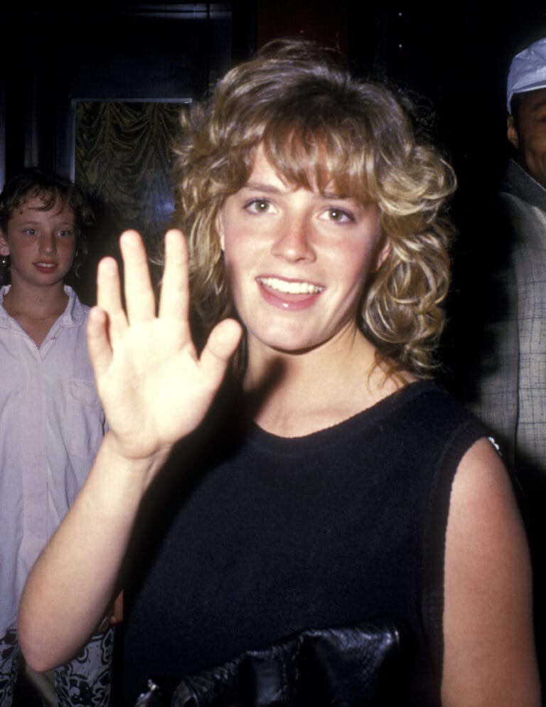 Elisabeth Shue: Wears a black sleeveless blouse, smiling and waving. She is in…