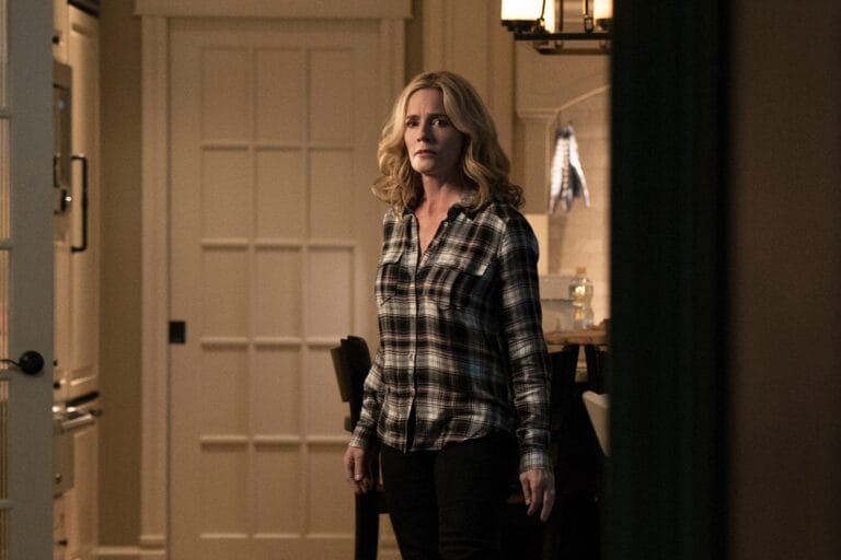 Elisabeth Shue: Wears a dark plaid shirt paired with tight black pants, standing upright in a home setting while…
