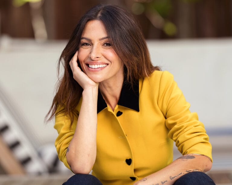 Sarah Shahi: Luce una blusa amarilla con cuello negro, posando sonriente mientras sostiene su rostro con…
