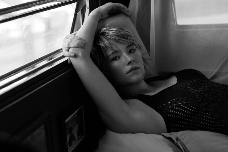 Haley Bennett: Luce un top negro de textura calada, recostada sobre una superficie suave mientras apoya…