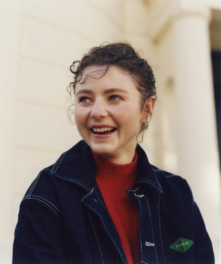 Thomasin McKenzie: Luce una chaqueta de mezclilla oscura sobre un suéter rojo de cuello alto, sonriendo…