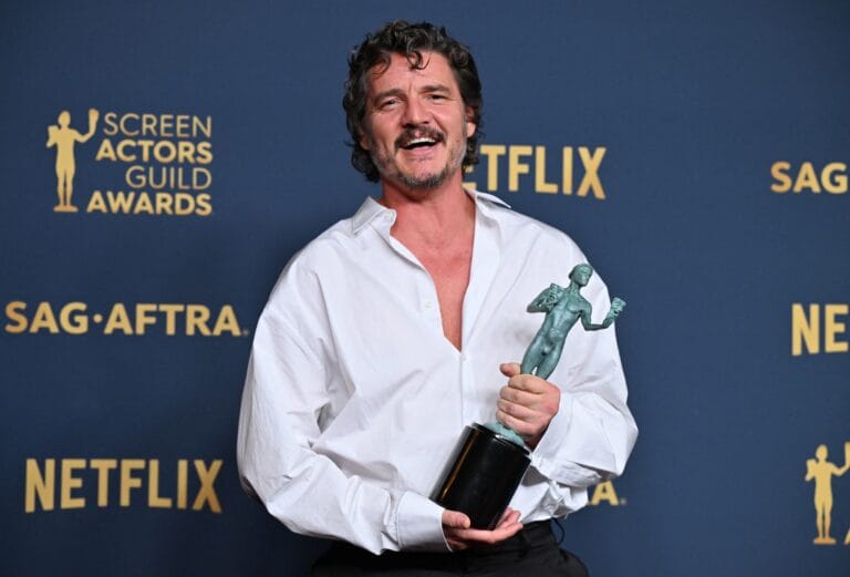 Pedro Pascal: Luce una camisa blanca de corte amplio, desabrochada en el pecho, mientras sostiene con…