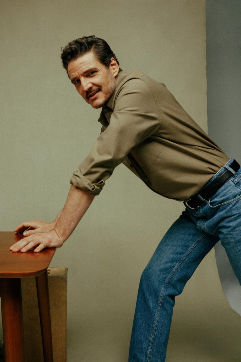 Pedro Pascal: Luce una camisa de tono tierra con las mangas arremangadas, apoyado sobre una mesa…