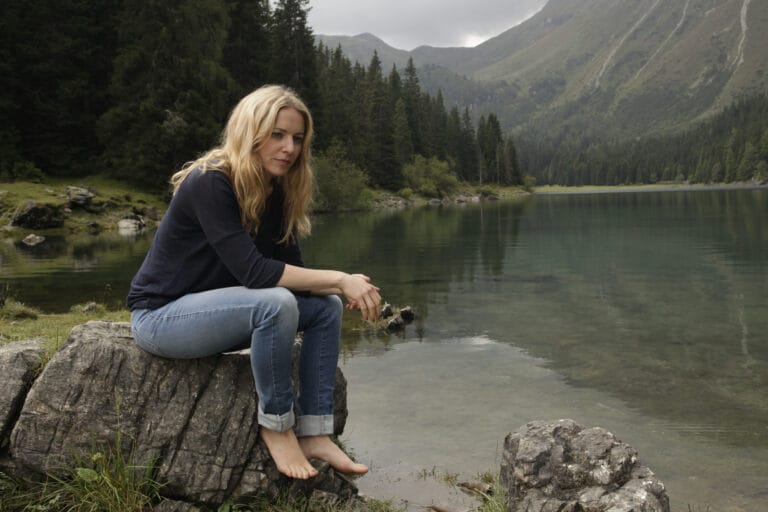 Diana Staehly: Está sentada sobre una roca junto a un lago sereno, con un suéter oscuro…