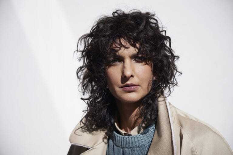Poorna Jagannathan: Luce un suéter azul con textura y una chaqueta beige sobre los hombros, creando…